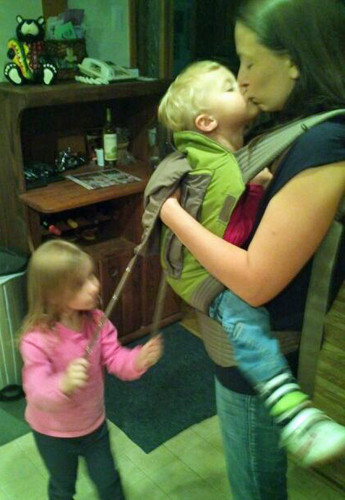 Chrystal getting a kiss from Kaylee while wearing her in an Onya Baby Carrier with Zoe pulling on the hood snaps