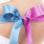 Pink and blue ribbon tied around the pregnant belly of a woman wearing white