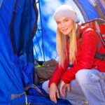 Young blond woman wearing beanie and red backpack squatting in front of her blue tent