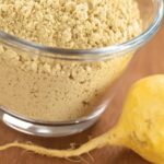 Close up of maca root powder in a clear glass bowl with maca root on the table next to it