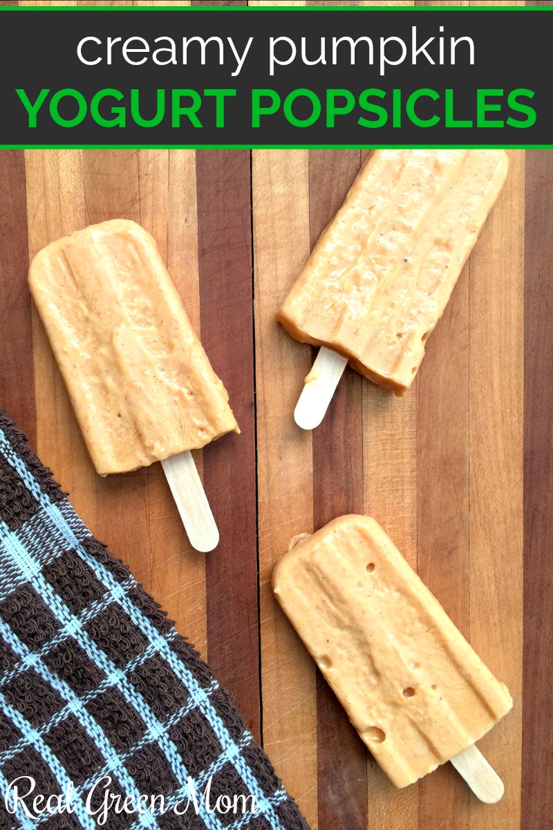 Three homemade pumpkin yogurt popsicles on a large wood cutting board next to a kitchen towel