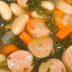 Close up of bowl of soup filled with slices of chicken sausage, white beans, kale and carrots
