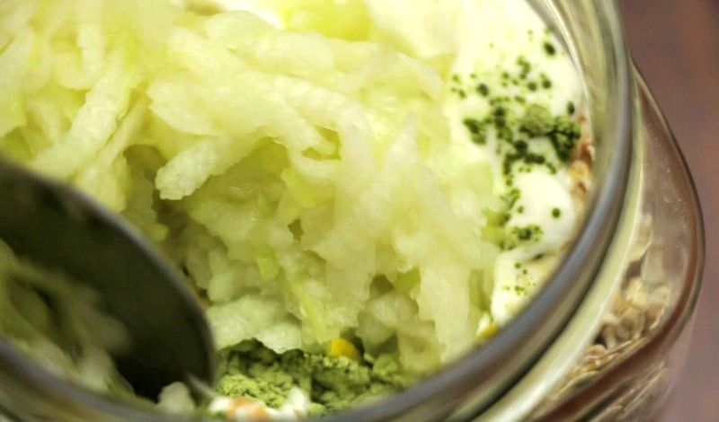 Oats, yogurt, matcha powder and grated apple being stirred together with a spoon in a jar