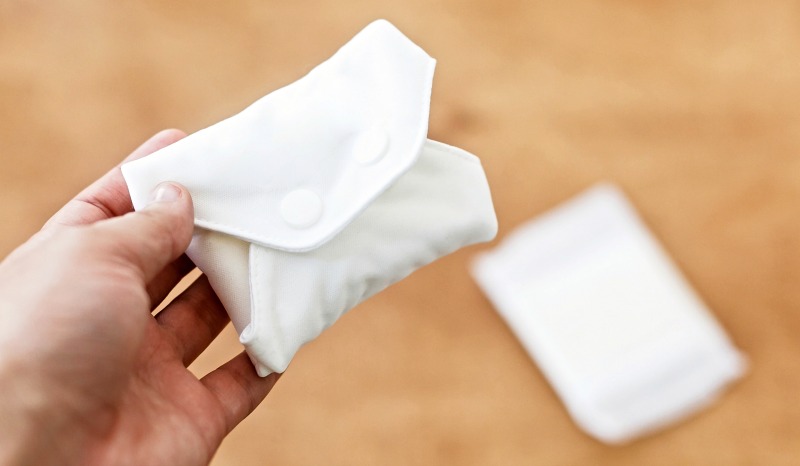 White reusable cloth pad snapped closed in a hand with a disposable pad on a table